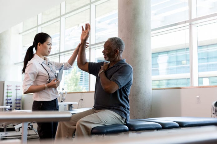 Confident female orthopedic doctor has a senior male patient lift his hand over his shoulder, testing his range of motion in his arm.